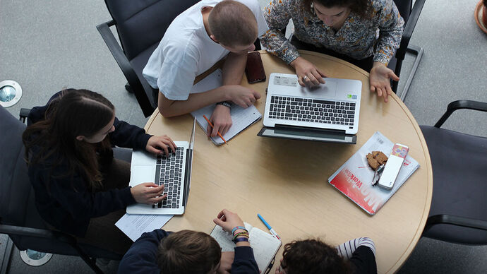 Blick von oben auf einen runden Tisch an dem vier Jugendliche sitzen, mit Schreibblöcken und Laptops