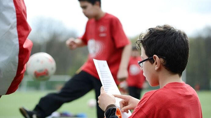 Ein Junge liest von einem Blatt etwas vor, im Hintergrund spielt ein anderer mit einem Ball